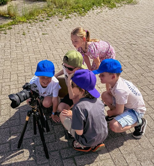 Vijf kinderen zitten in een kring om de camera op statief en kijken samen naar het schermpje